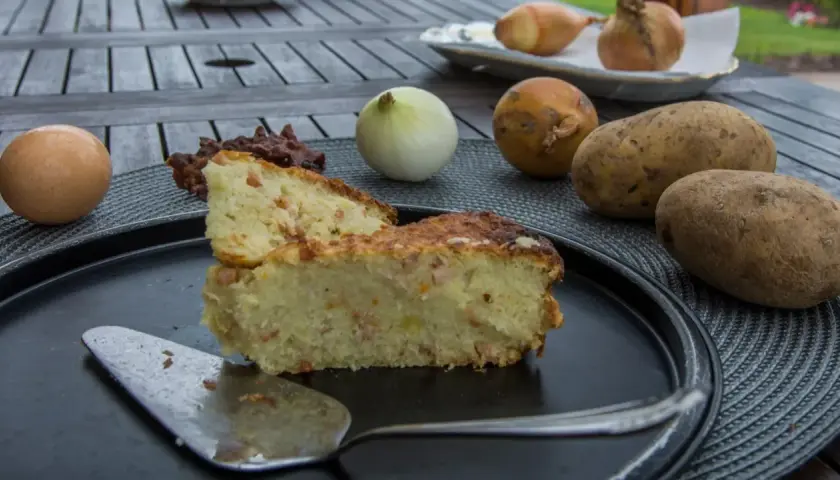 Babka ziemniaczana przepis babci: złocista, pachnąca babka ziemniaczana z boczkiem, obok surowe ziemniaki, cebula i jajko.