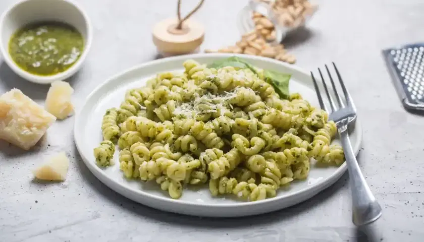 Pyszny makaron z pesto ze słoika, posypany parmezanem. Obok miseczka z zielonym sosem i kawałki sera.