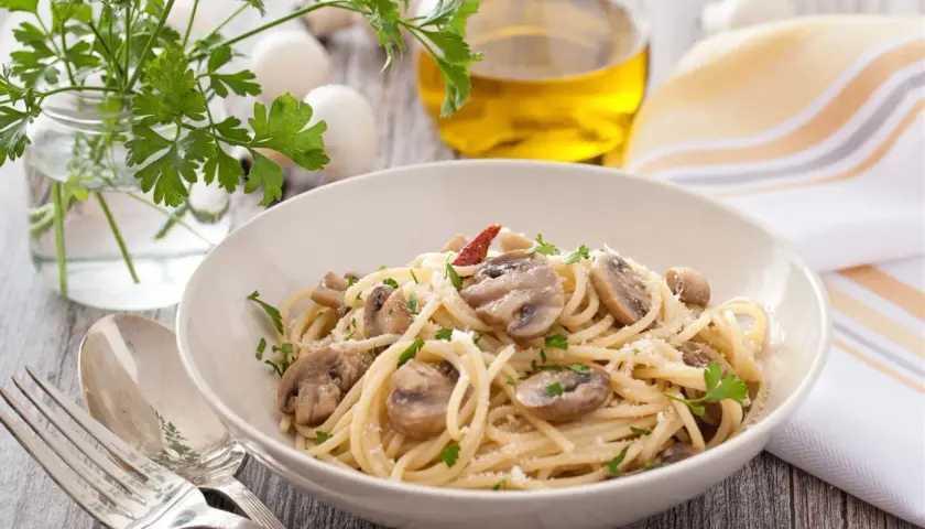 Talerz spaghetti z pieczarkami, posypany parmezanem i natką pietruszki. Obok sztućce, oliwa i zioła.