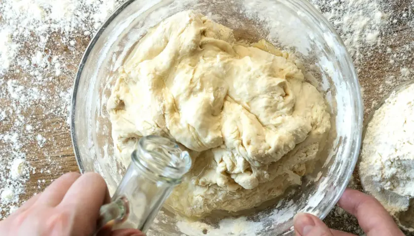 Dłoń dodaje olej do ciasta na roti w szklanej misce, otoczonej mąką.