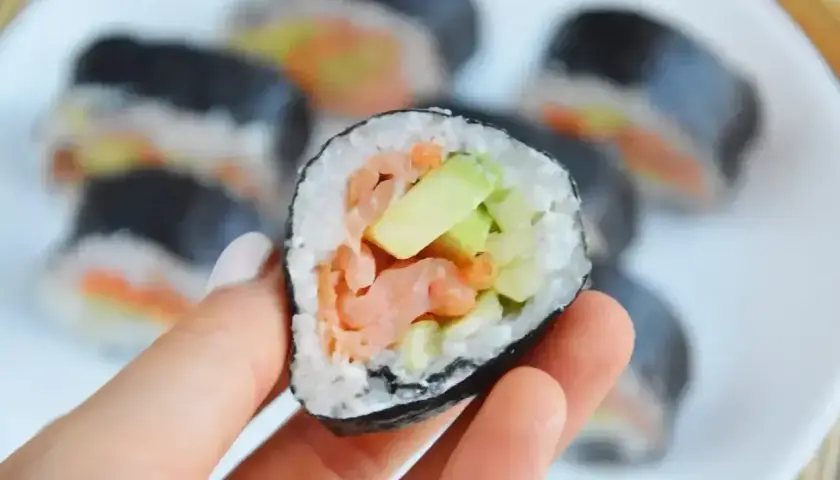 Ręka trzyma kawałek futomaka z łososiem i awokado. Na talerzu widać więcej sushi.