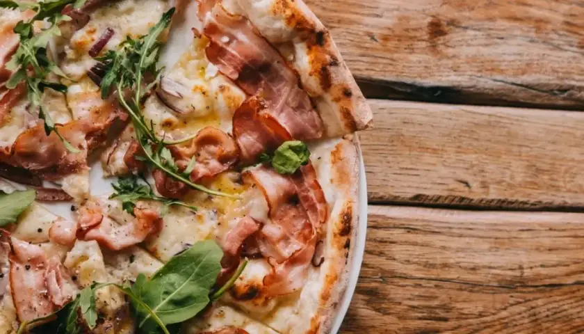 Pyszna pizza carbonara z chrupiącym boczkiem i rukolą, podana na drewnianym blacie.