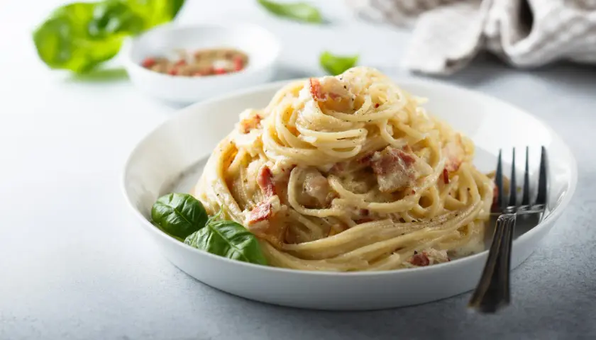 Pyszna carbonara z boczkiem, podana na białym talerzu z listkami bazylii i widelcem.
