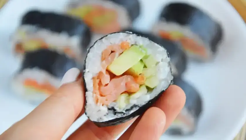 Oto futomaki co to: ręka trzyma kawałek sushi z łososiem i awokado. W tle talerz z innymi rolkami.