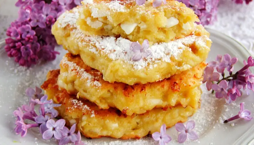 Stosik puszystych placuszków z serka wiejskiego i banana, posypanych cukrem pudrem i ozdobionych bzem.
